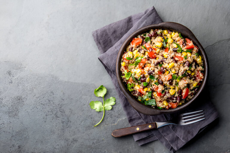 A colorful quinoa salad with black beans, corn, and fresh vegetables in a dark bowl on a gray surface.