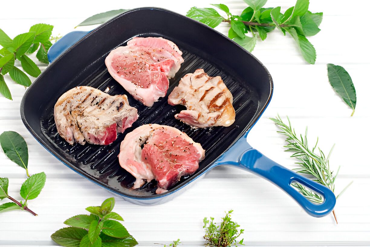 Grilled meat chops in a blue grill pan surrounded by fresh herbs