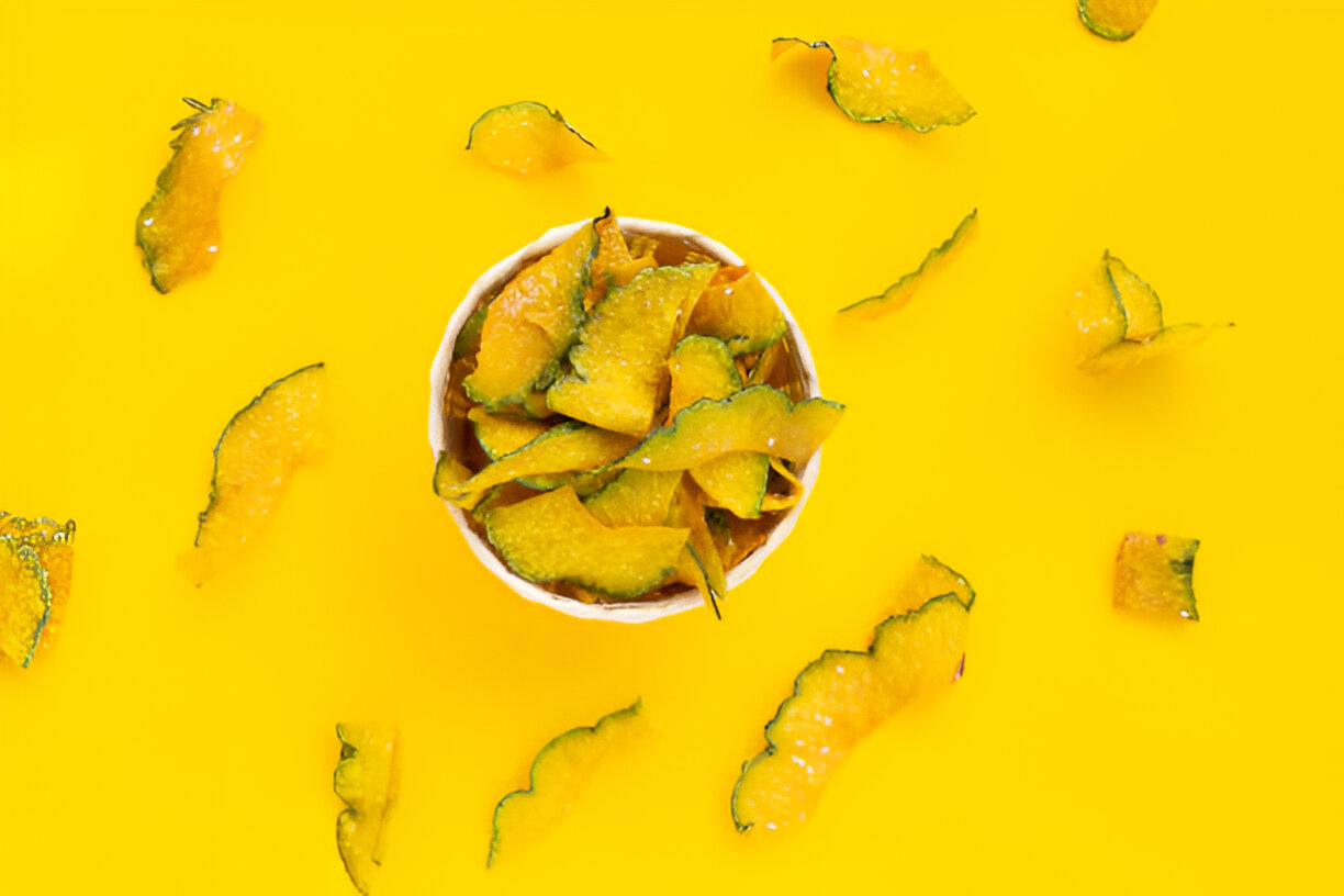 A bowl of crispy, vibrant yellow pumpkin chips surrounded by scattered pumpkin peel on a bright yellow background.
