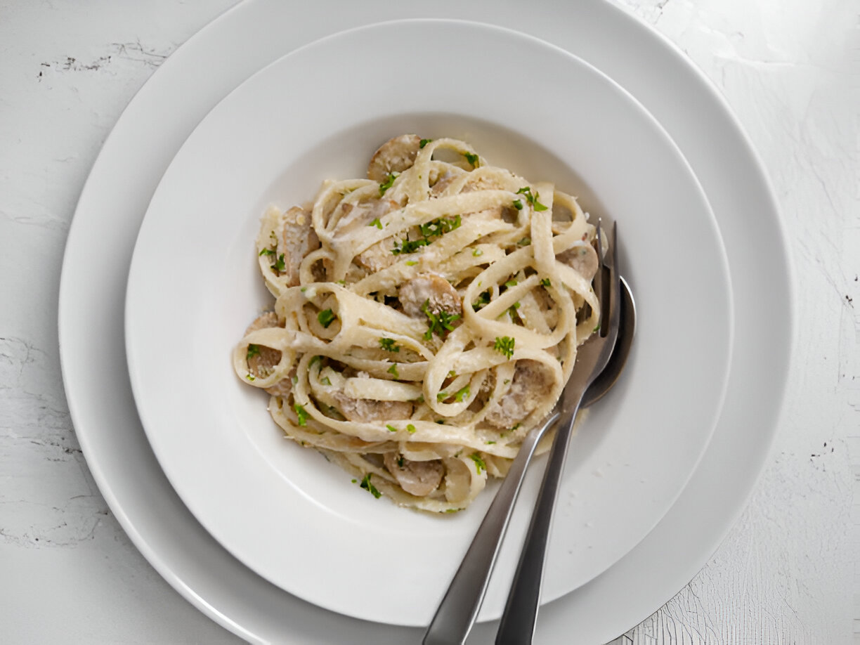 A bowl of fettuccine pasta with creamy sauce and herbs served in a white dish on a textured surface.