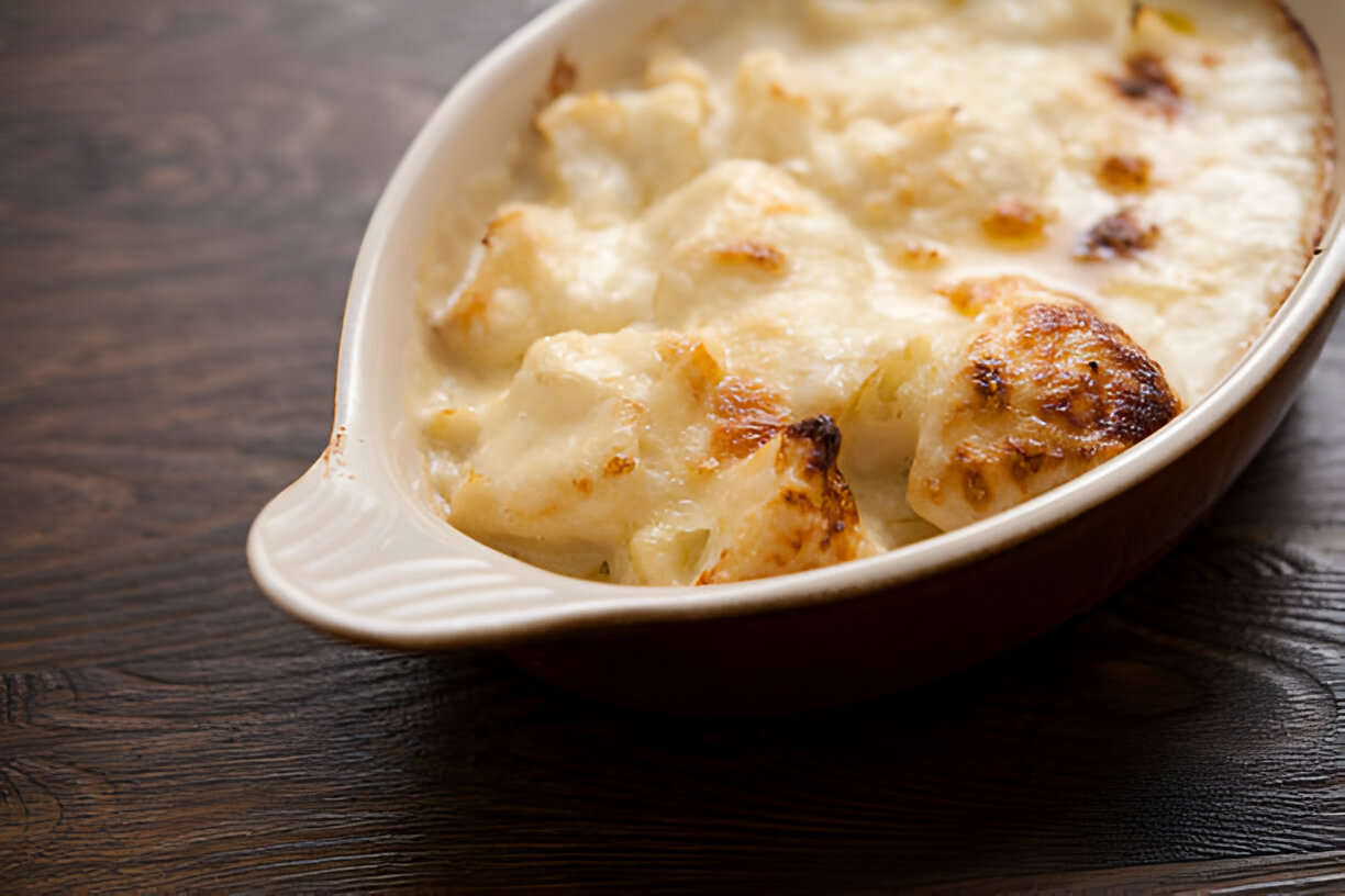 A baking dish filled with creamy, golden-brown cauliflower gratin sitting on a wooden surface.