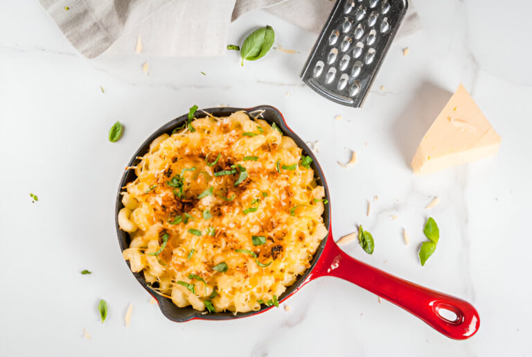 A delicious serving of macaroni and cheese in a red skillet, garnished with herbs and accompanied by a block of cheese and a grater.
