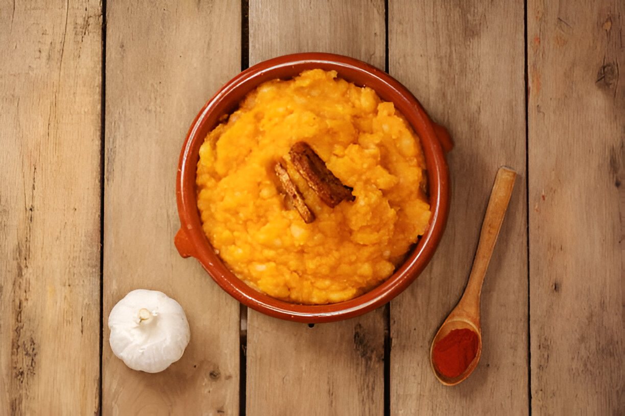 A bowl of orange mashed food topped with a piece of cinnamon in a clay dish, accompanied by a garlic bulb and a wooden spoon with red spice on a wooden surface.