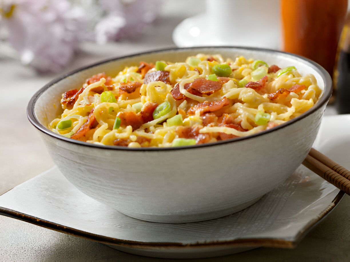 A bowl of savory noodle dish with green onions and bacon on a white background.