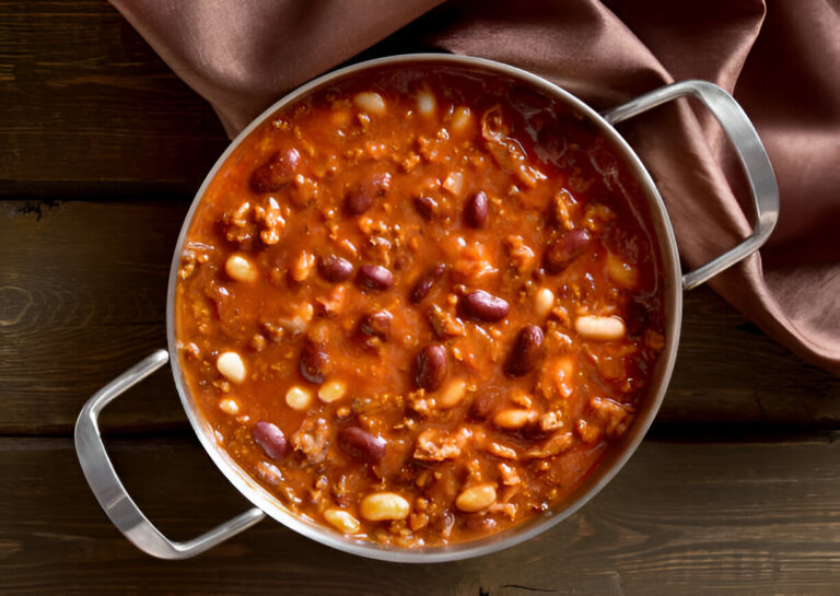 A pot of hearty chili with kidney beans and other ingredients, resting on a wooden surface next to a brown cloth.