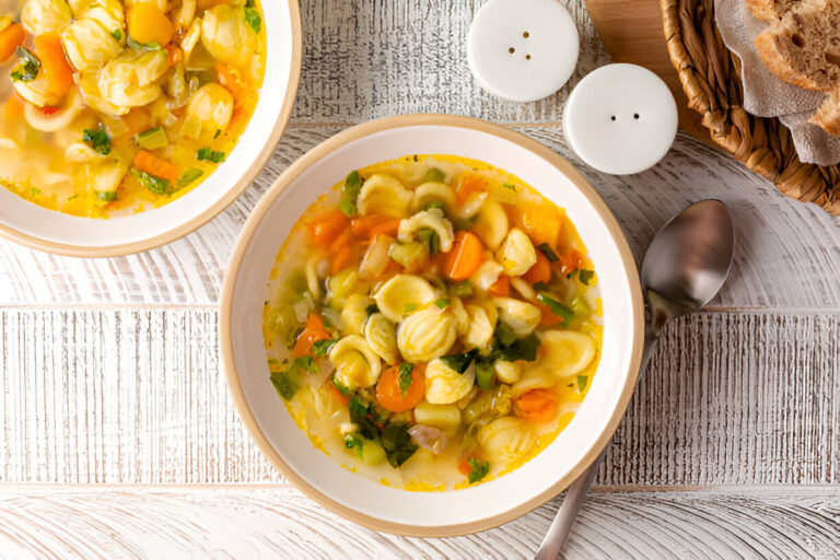 A bowl of colorful vegetable soup with pasta on a rustic wooden surface.