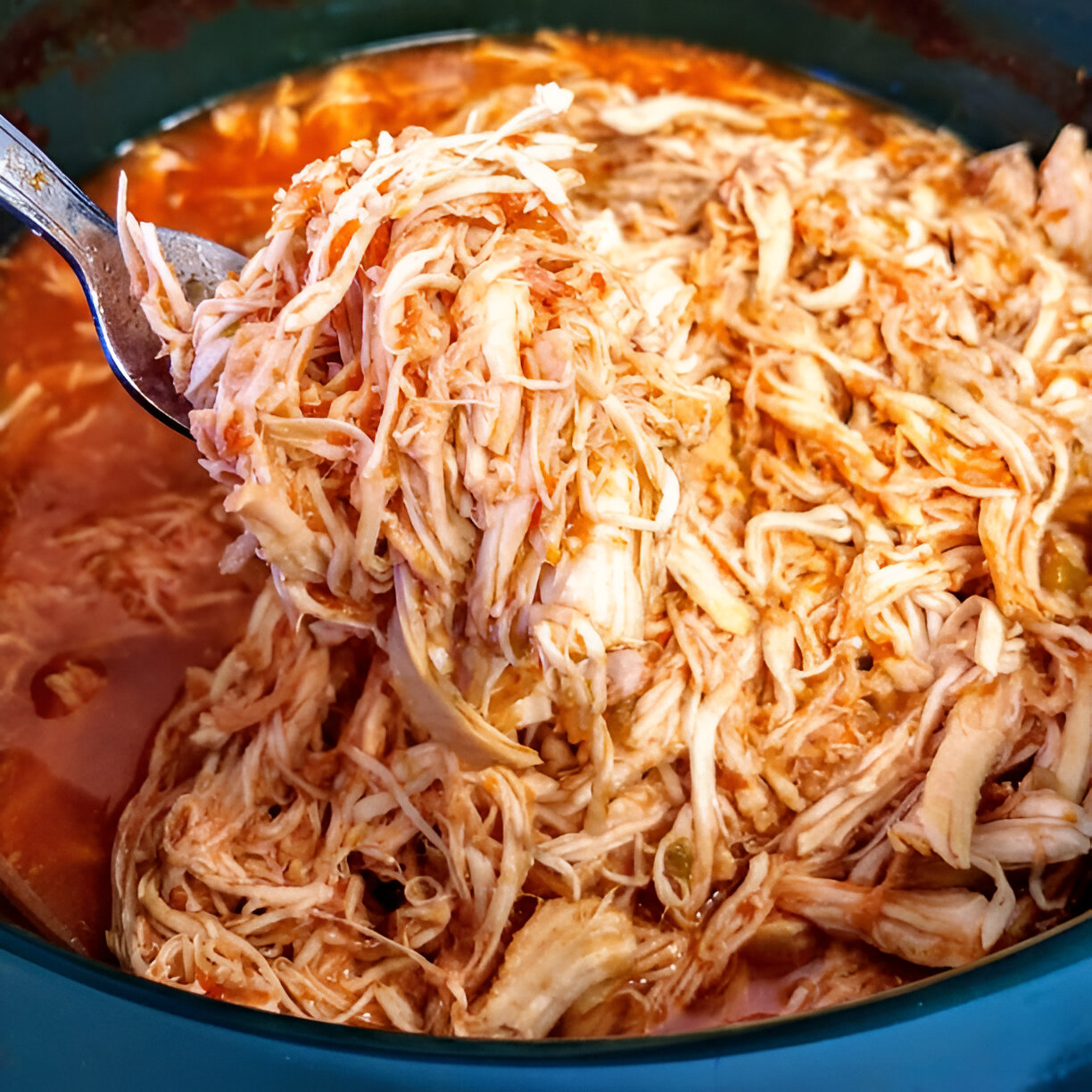 A close-up of shredded chicken in a savory sauce, being lifted from a pot with a spoon.