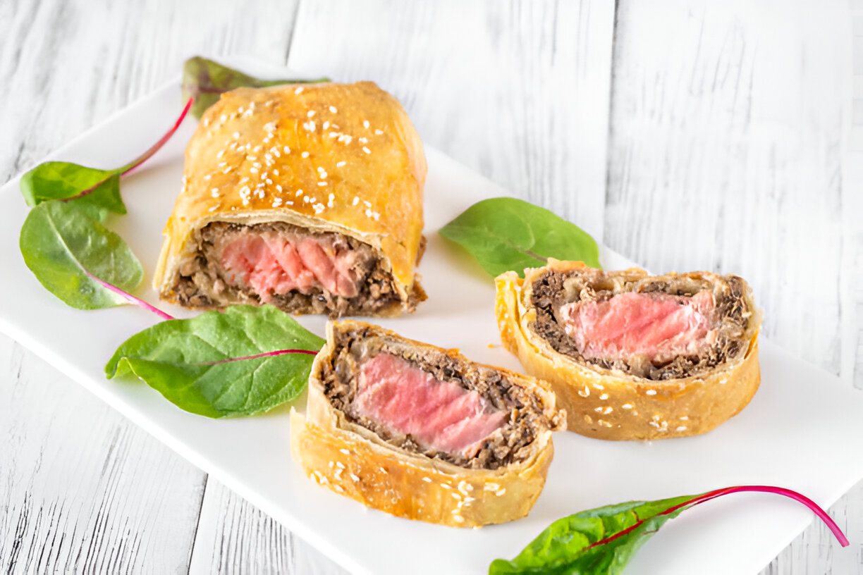 A plate of sliced beef Wellington, showcasing the flaky pastry and pink beef filling, garnished with fresh greens.