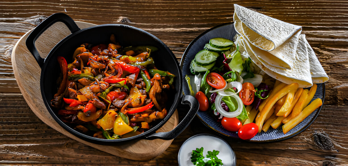 A colorful dish served in a black cast iron pot with mixed vegetables and meat, accompanied by a side of salad, fries, tortillas, and a small bowl of sauce.