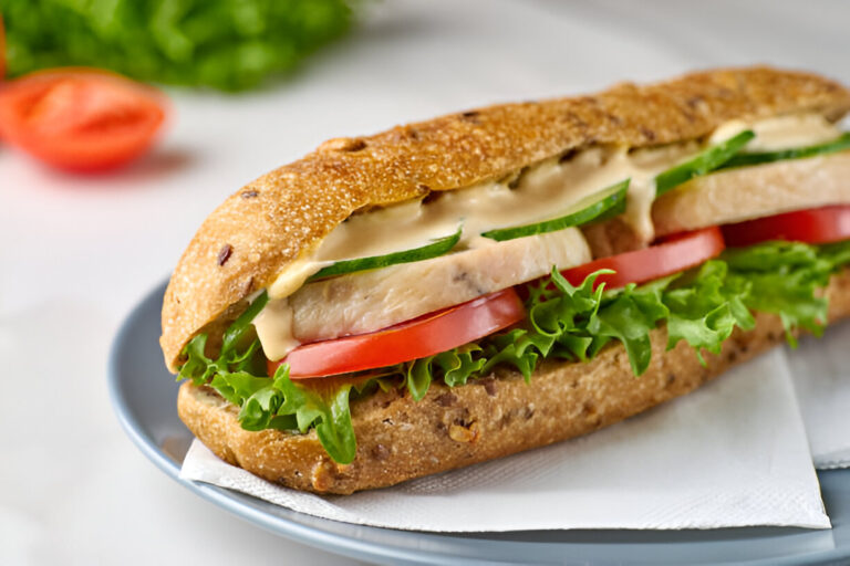 A fresh sandwich on a plate, featuring whole grain bread filled with lettuce, tomato, cucumber, and slices of chicken, topped with a creamy sauce.