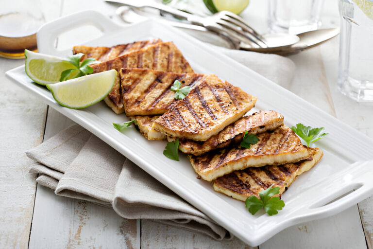 Grilled slices of tofu garnished with lime wedges and fresh herbs on a white platter.