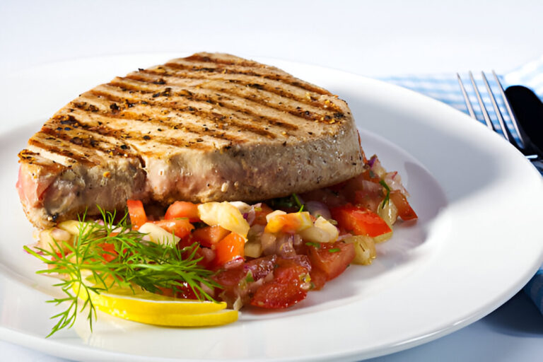 A grilled tuna steak served on a white plate with a colorful tomato salad and a slice of lemon