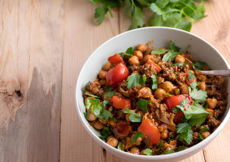 A bowl of chickpea and ground meat dish topped with fresh parsley and red bell peppers, placed on a wooden table.