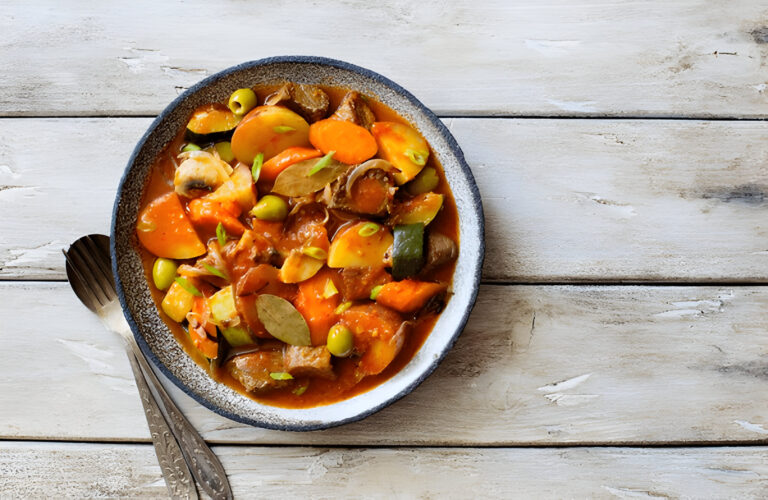 A bowl of colorful vegetable stew with carrots, potatoes, olives, and herbs on a rustic wooden table.