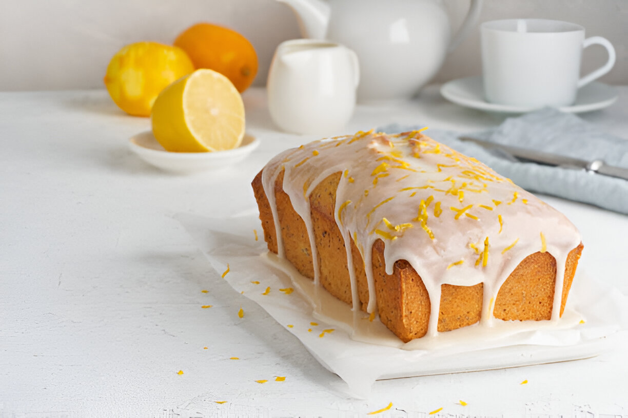 A freshly baked lemon loaf cake with icing and lemon zest on top, arranged on a white surface with lemons in the background.