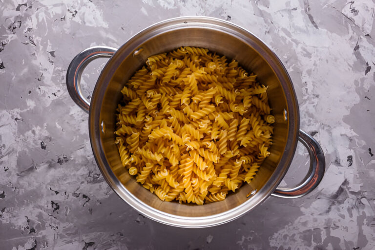 A stainless steel pot filled with uncooked fusilli pasta on a textured gray background.