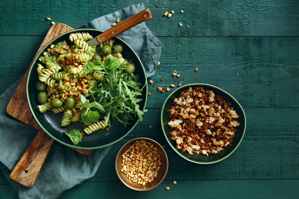 A vibrant dish featuring green pasta, fresh arugula, olives, and a scattering of ingredients on a wooden table.