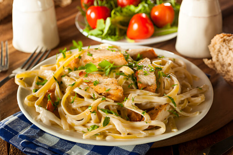 A plate of creamy fettuccine pasta with grilled chicken, garnished with parsley, alongside a side salad with cherry tomatoes.