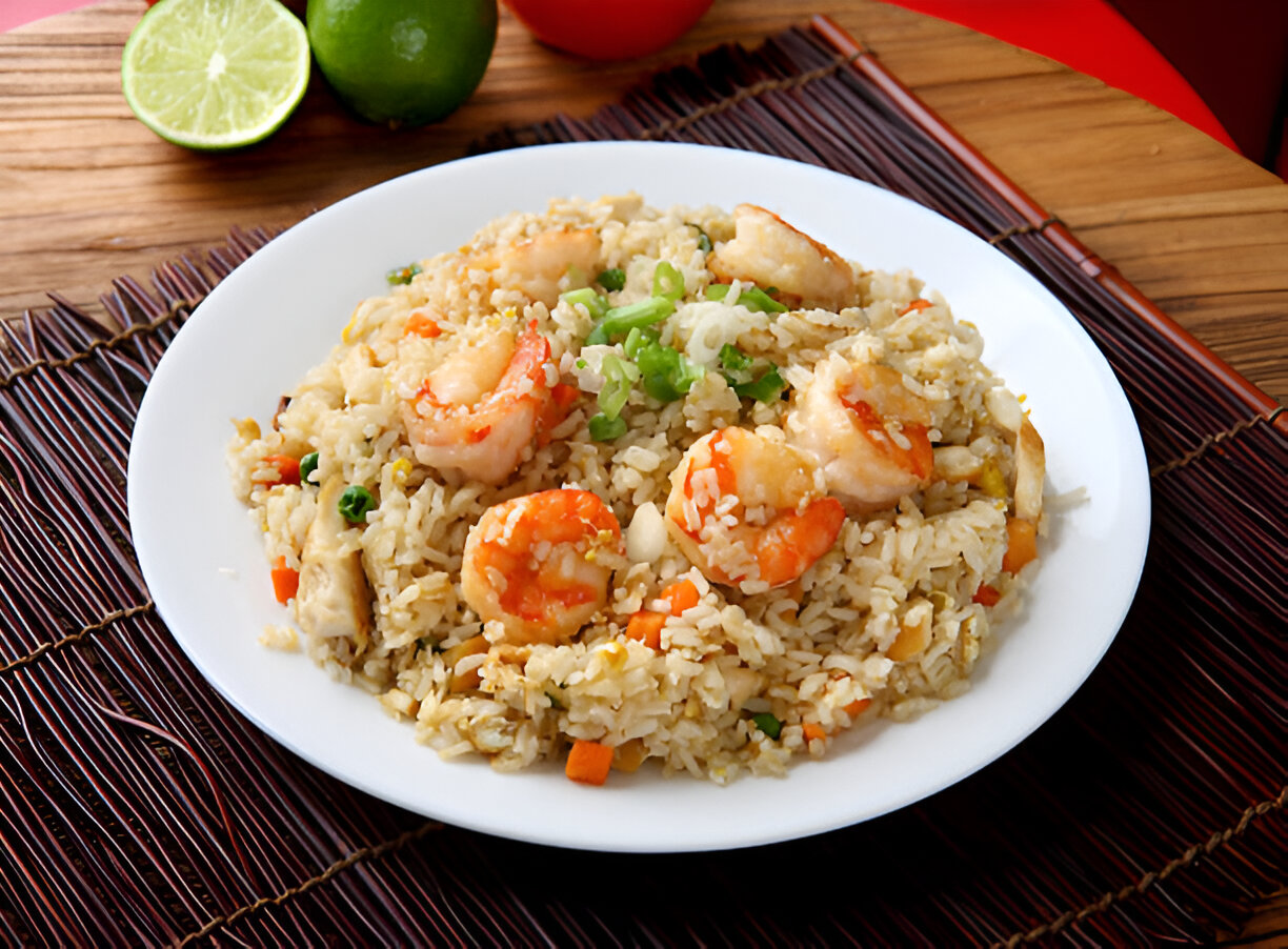 A plate of shrimp fried rice garnished with green onions, served with lime and tomatoes in the background.