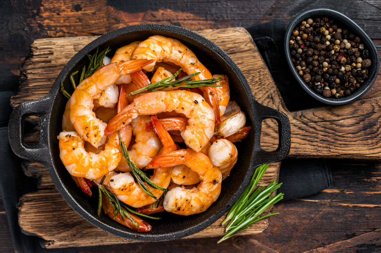 A cast iron skillet filled with cooked shrimp garnished with herbs, next to a small bowl of mixed black peppercorns.