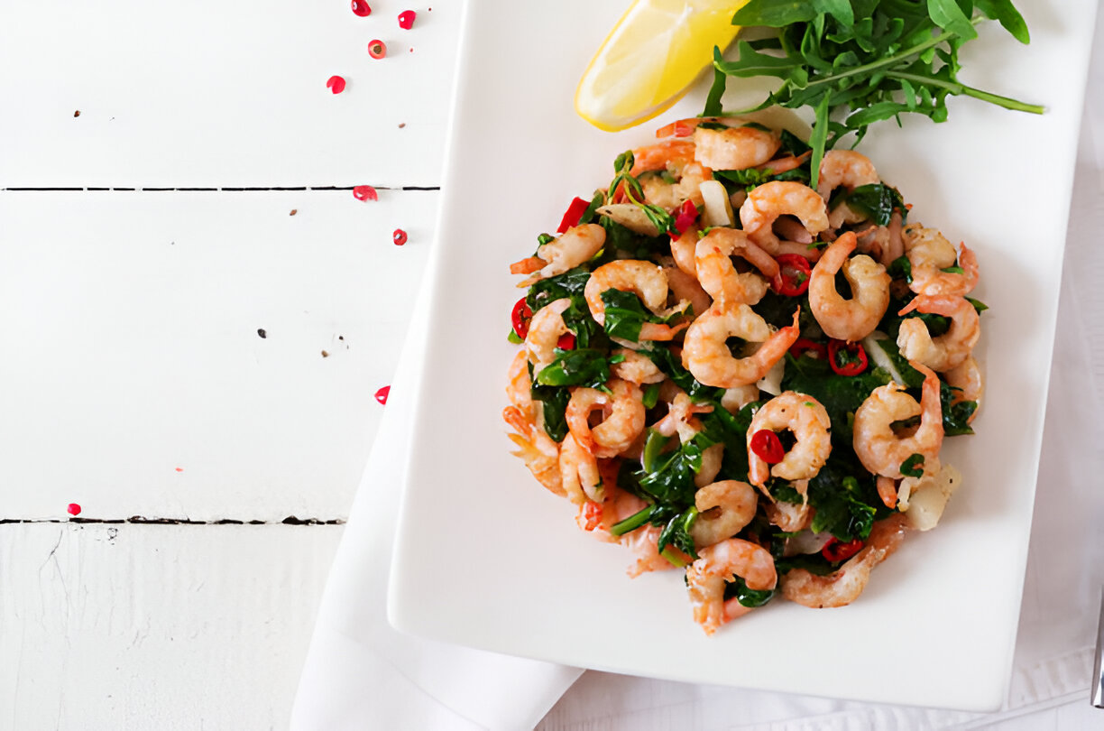 A plate of shrimp salad with mixed greens and chili peppers, garnished with lemon and fresh arugula.