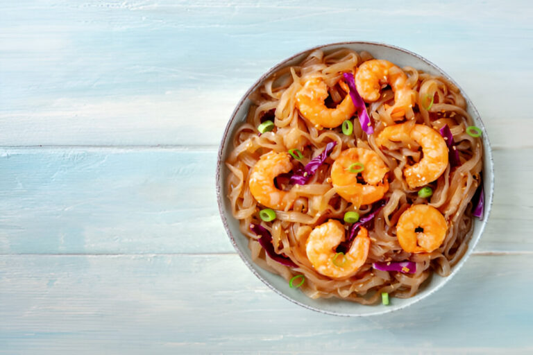 A delicious bowl of shrimp noodles with purple cabbage and green onions on a light blue wooden table.