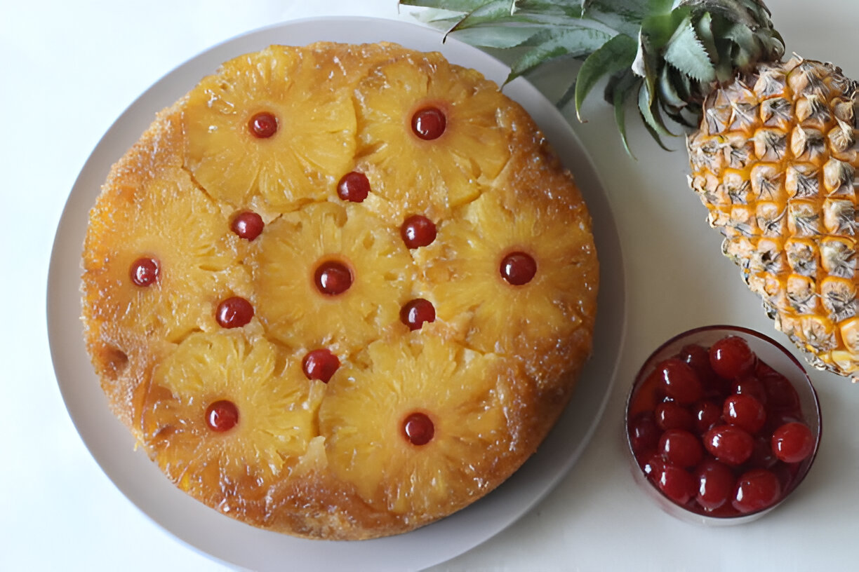 A delicious pineapple upside-down cake garnished with cherries, served on a white plate next to a fresh pineapple and a bowl of maraschino cherries.